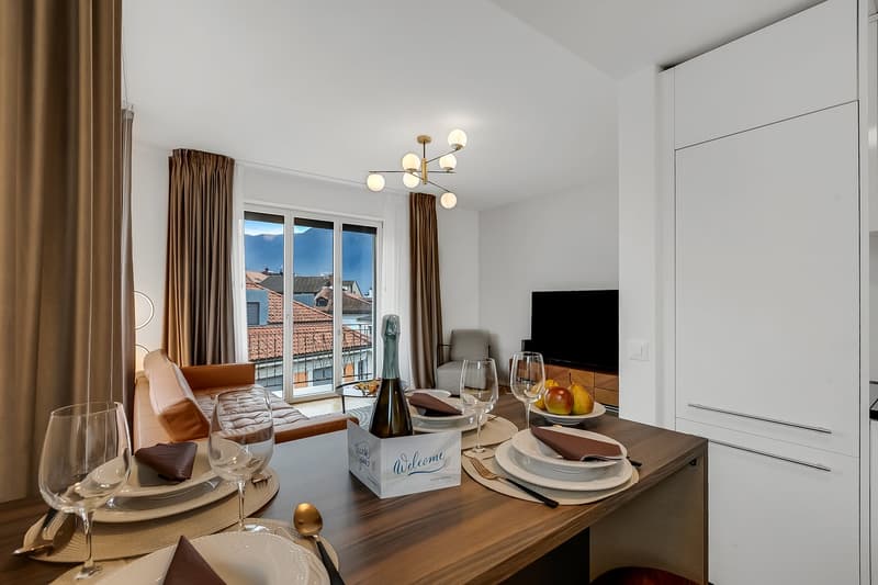 Dining area with a table, chairs, and a bottle of wine. A couch, a TV, and a chandelier are in the background. A window with a view of the city is on the wall.
