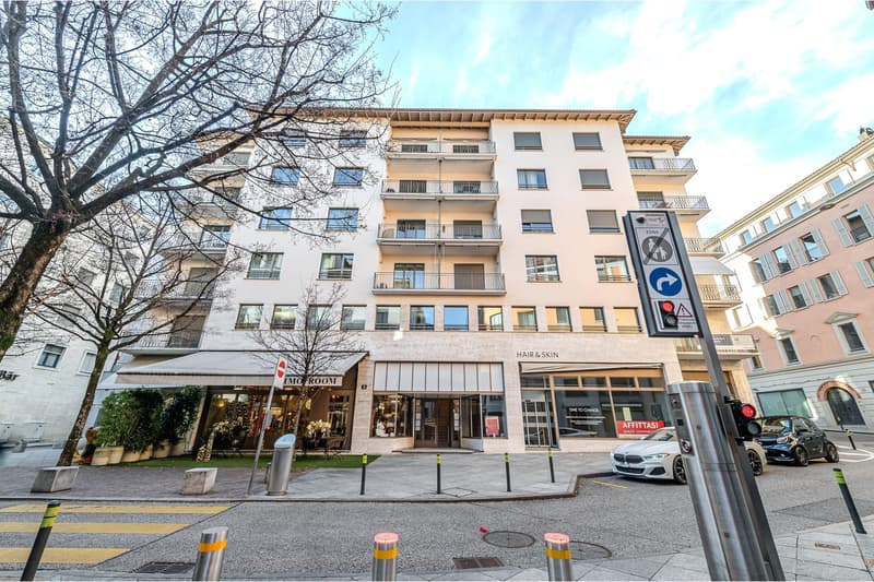 Modern multi-story building with glass windows, balconies, and a ground floor with shops.