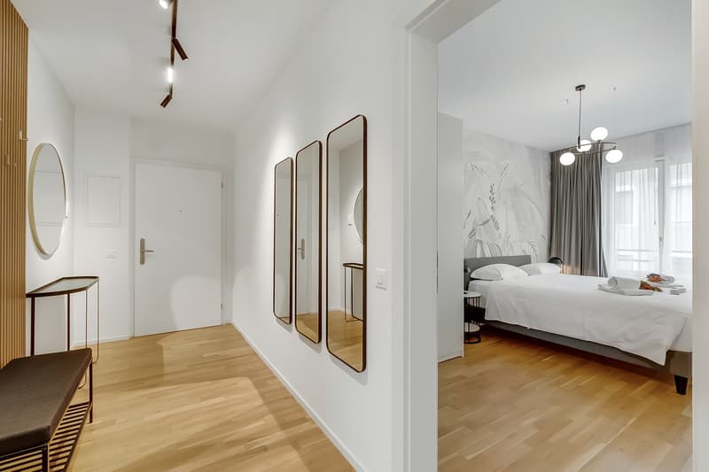 Modern bedroom with white walls, wooden floor, large bed, white bedding, three mirrors, wooden bench, and a small table with a lamp