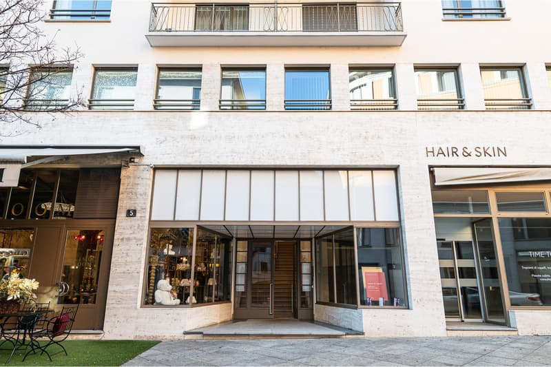 White building with glass windows and doors, entrance with glass doors, sign reading Hair & Skin, staircase inside, shop windows, outdoor seating area with table and chairs
