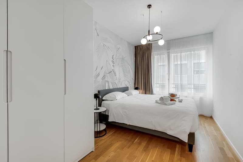 modern bedroom with white walls, wooden floor, large bed, nightstands, lamps, large windows, white curtains