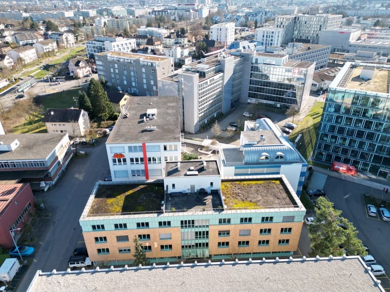 Aerial view of a group of buildings, with various sizes and architectural styles, surrounded by greenery and a parking lot.