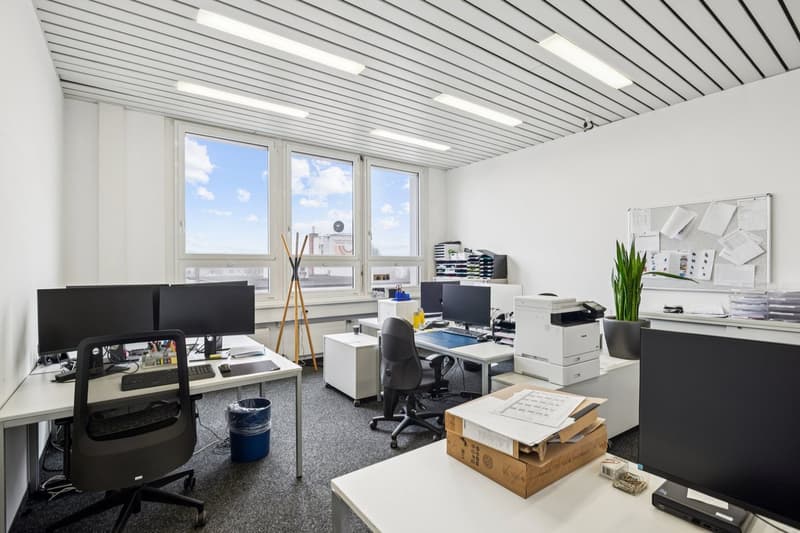 modern office with white walls, white desks, black office chairs, computer monitors, printer, potted plant, bulletin board, trash bin