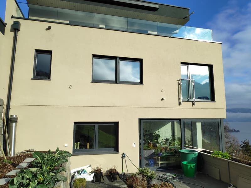 2 story modern house with beige exterior, large windows, glass balcony, and a garden area with plants and gardening equipment visible.