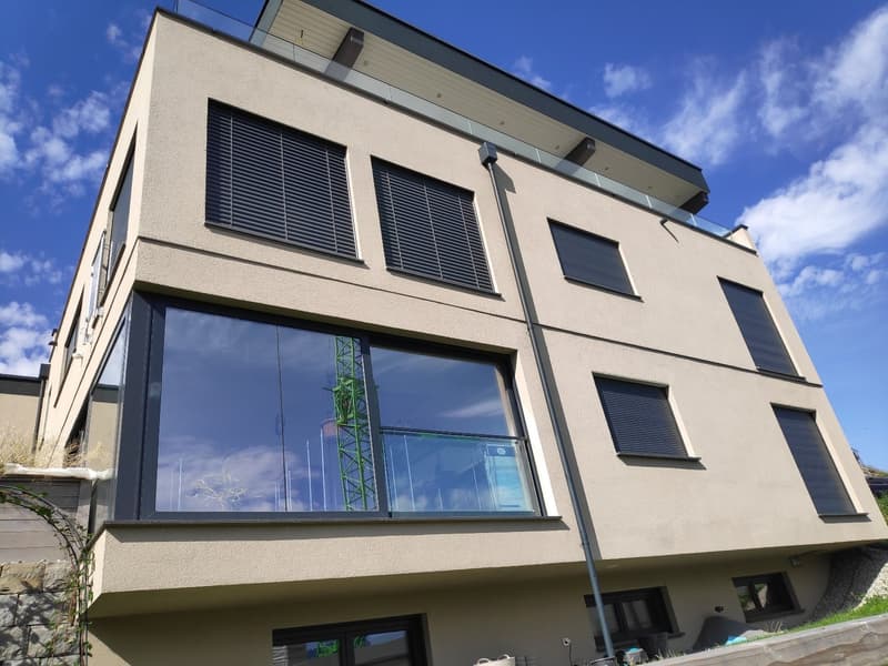 3-story modern building with a beige exterior, large windows, and a balcony on the top floor. The building has a sleek, contemporary design with black window frames and solar panels on the roof.