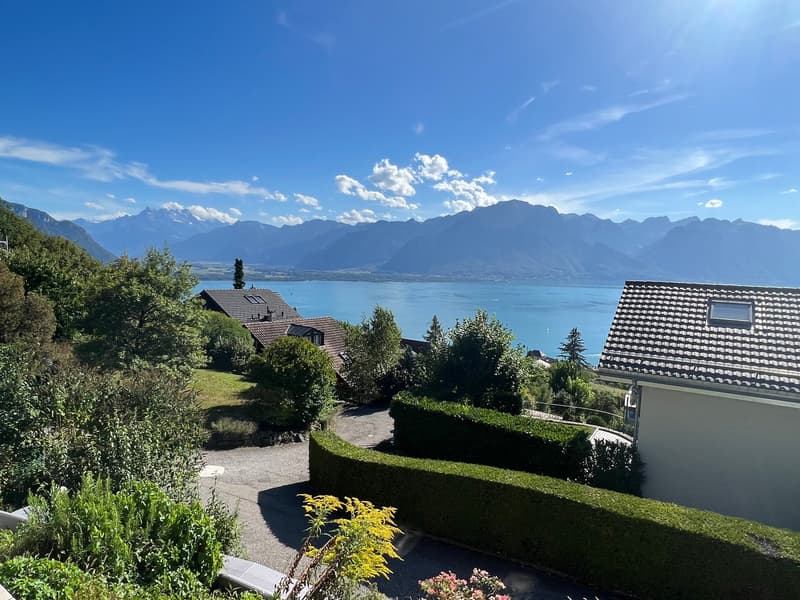 The image shows a scenic view of a lakeside town surrounded by mountains. There are several houses with red-tiled roofs nestled among lush greenery, including trees and hedges. The lake in the foreground reflects the blue sky and clouds above, creating a