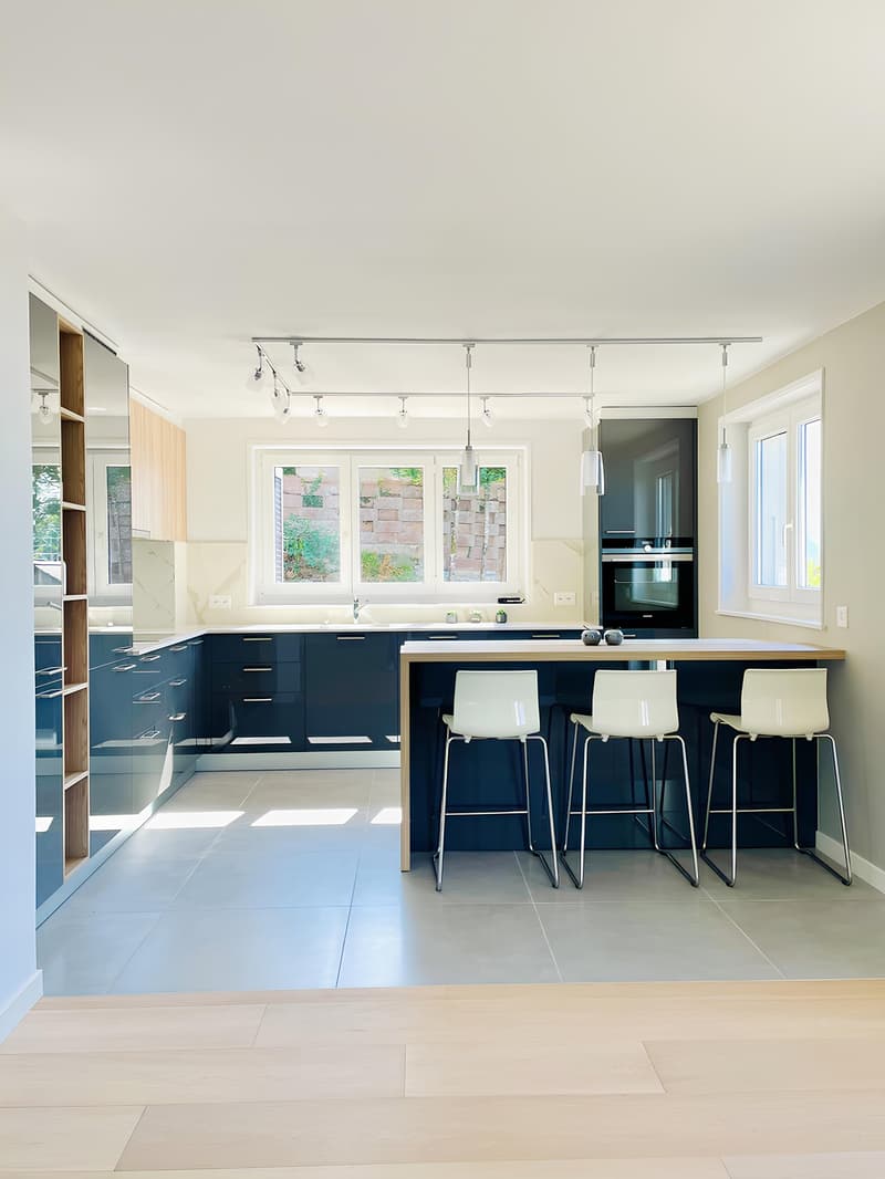 Modern kitchen with navy blue cabinets, wooden countertops, white bar stools, and large windows overlooking a garden. The kitchen features a built-in dishwasher and track lighting.