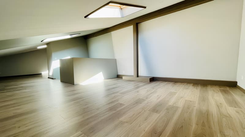 Spacious living room with hardwood flooring, exposed wooden beams, and a recessed lighting fixture. The room has a minimalist design with a fireplace visible on the right side.