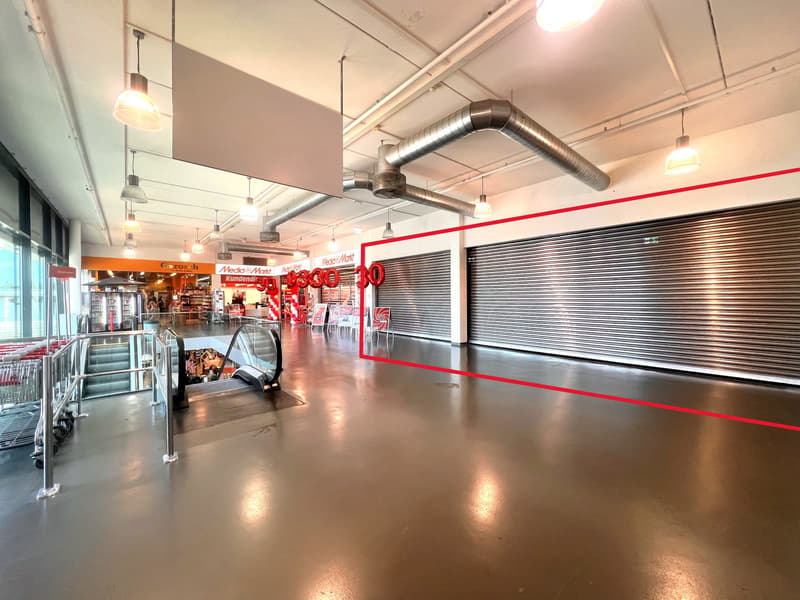 Open space with a shopping mall in the background. Empty, grey floor, and two rows of silver roll-up shutters. There is a double stair in the back.