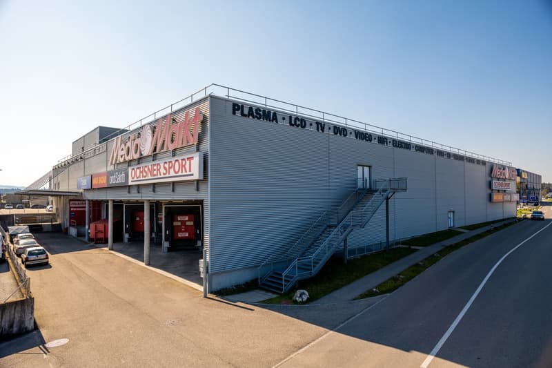 large gray building with 'Media Markt' sign, multiple stores below, stairs on right, red and white signs, cars parked, street in front