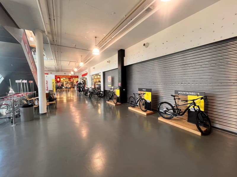Bicycle showroom with bicycles on display, grey floor, white and silver walls, silver bicycle rack, and multiple shops visible in the background.