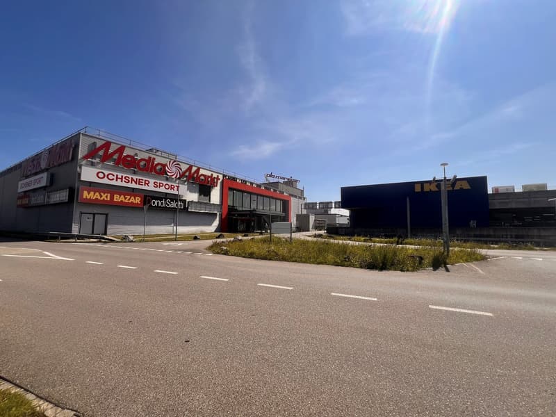 A commercial building with white walls, red accents, and a prominent 'Media Markt' sign. It is a large structure with glass windows and a modern design. Nearby, there is a grassy area and a sign for 'Maxi Bazar'. On the right, another building with a blue