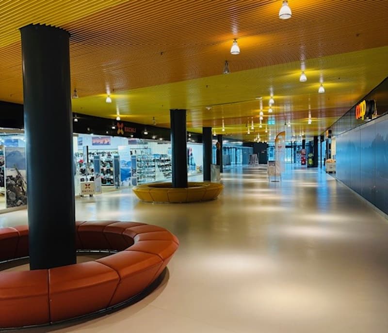 Empty mall with yellow ceiling, black columns, orange seating, white floor, shops on the side