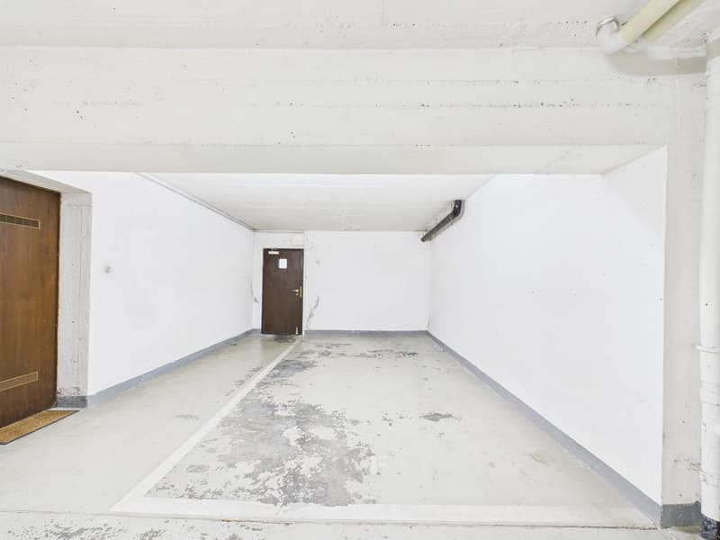 Empty parking garage with white walls, concrete floor, two wooden doors, and a pipe on the ceiling.