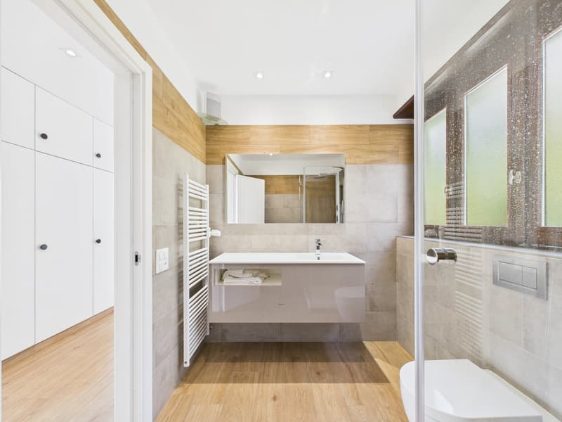 modern bathroom with white tiles, wooden floor, white vanity, mirror, glass shower enclosure, white toilet, towel rack
