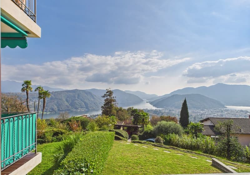 House with balcony, green railings, overlooking a lake and mountains, lush green garden, palm trees, and a distant town.