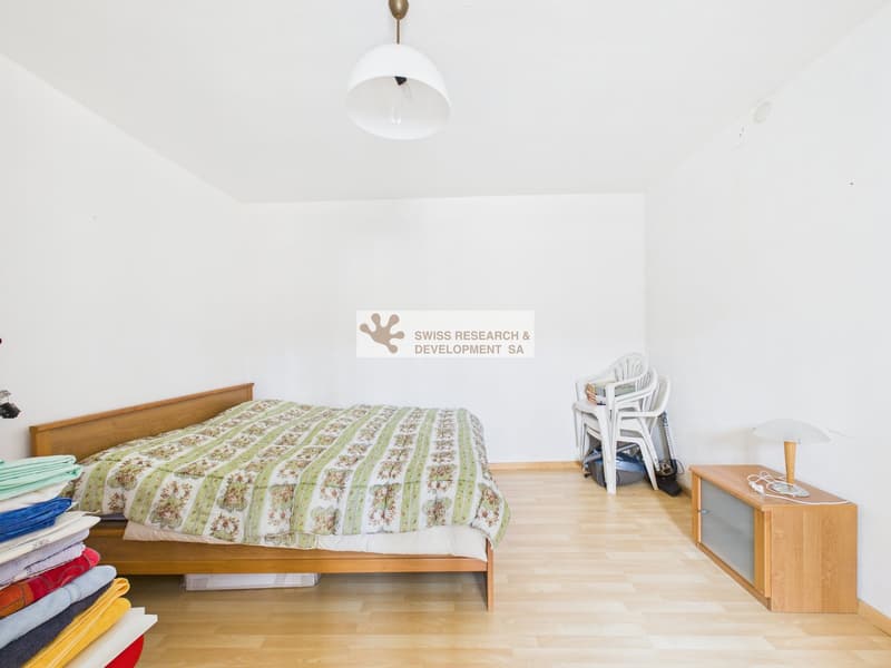 Bedroom with bed, lamp, wooden floor, white walls, nightstand with lamp, stack of towels, stack of clothes, chair, and table
