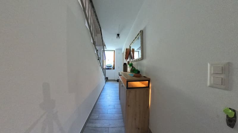 Empty hallway with white walls, wooden floor, a wooden cabinet with a lit up display, a mirror, and a switchboard.