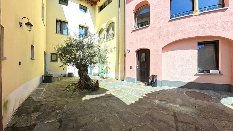 Two buildings with yellow and pink walls, stone paved courtyard, tree, lamp, trash bin, door
