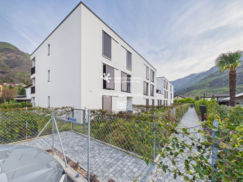Modern white building, multiple windows, metal fence, paved walkway, greenery, mountains in the background