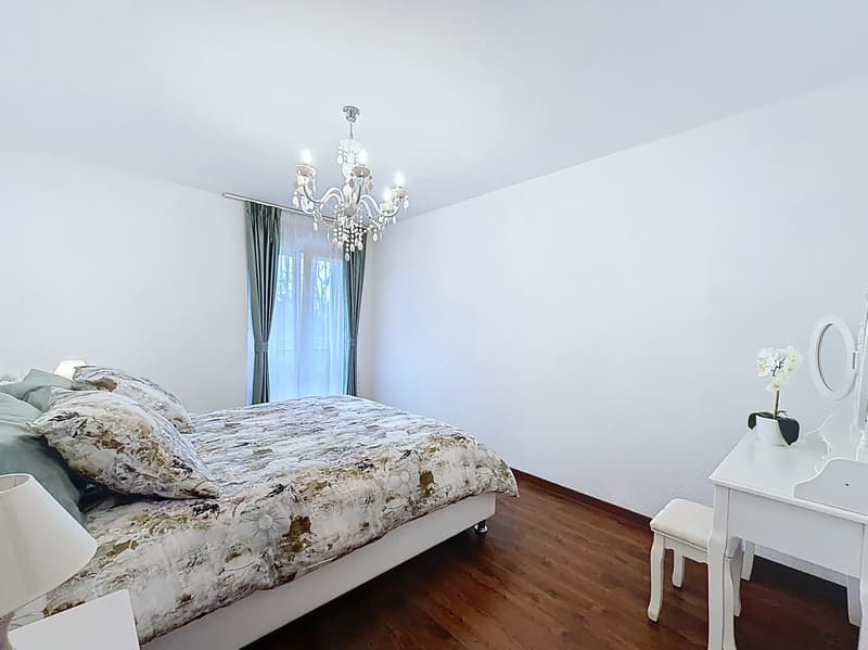 Bedroom with a bed, pillows, a chandelier, a desk, a mirror, a stool, a lamp, and a window with curtains.