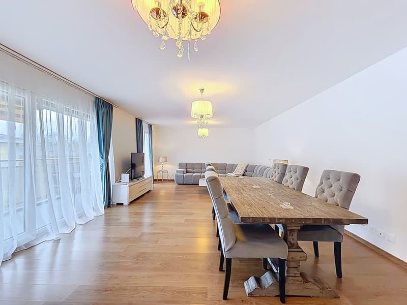 Dining room with wooden table, gray chairs, chandeliers, white walls, large windows, white curtains, TV, cabinet, couch, lamp