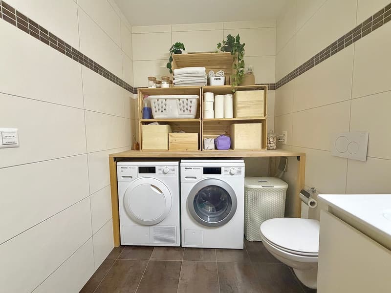 small laundry room, with white washer, dryer, a shelf above the washer and dryer, toilet, trash can