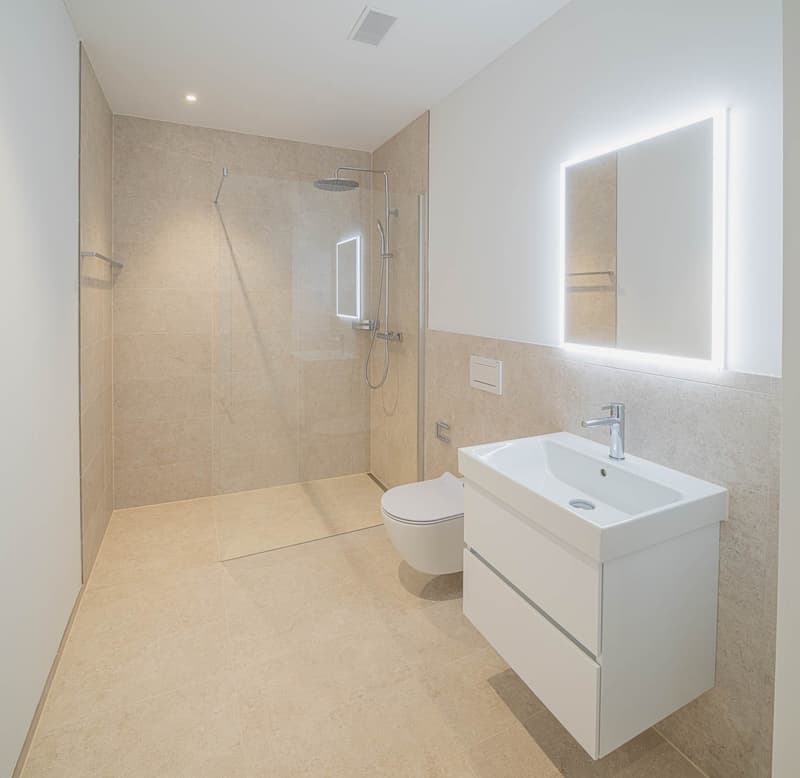 Spacious bathroom with beige tile walls and floors, walk-in shower with glass enclosure, vanity with sink, and toilet. The bathroom also features a lighted mirror and appears to have a private washing machine and dishwasher installed.