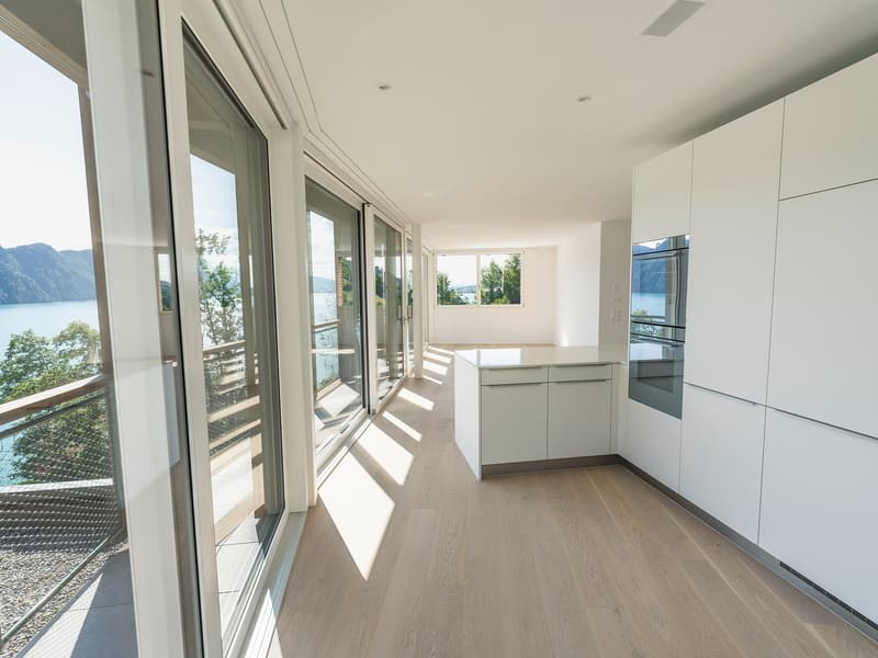 Spacious open-plan living area with large windows and sliding glass doors leading to a balcony or terrace, offering a scenic view of the lake and mountains. The room has a modern, minimalist design with white walls, hardwood floors, and built-in kitchen c