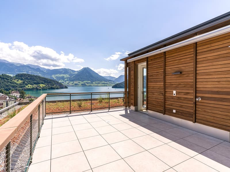 2-story wooden house with a large balcony overlooking a lake surrounded by mountains. The balcony has a tiled floor and metal railing. The house has a modern design with large windows and a scenic view of the lake and mountains.