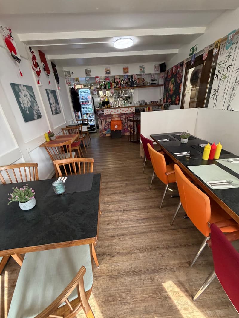 Wooden floor, white walls, tables and chairs, refrigerator, counter with bottles, wall decorations, ceiling light
