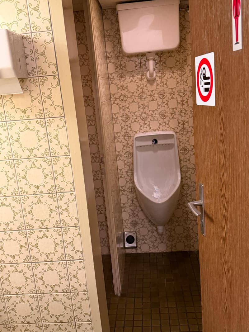 A small bathroom with a urinal and a toilet, tiled walls and floor, wooden door, and a sign on the door