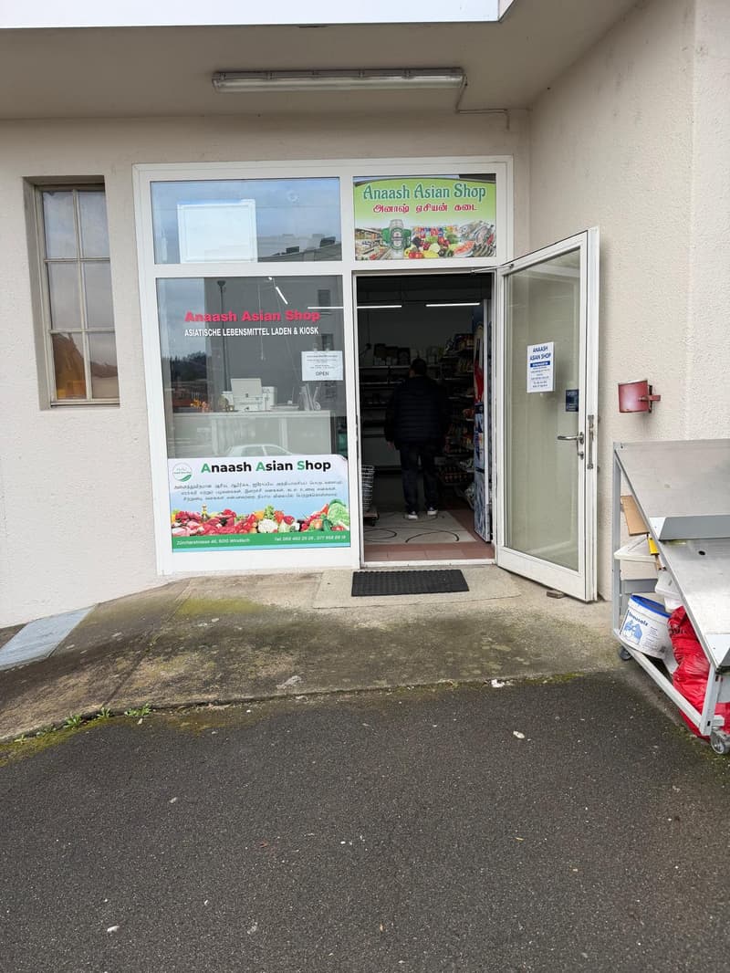 Storefront of a shop with glass door and windows, open door, person inside, signboard, shelves with products, trash bin, and a trolley with buckets