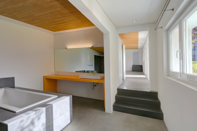 White bathtub, rectangular sink, rectangular mirror, wooden ceiling, staircase, large window, concrete floor