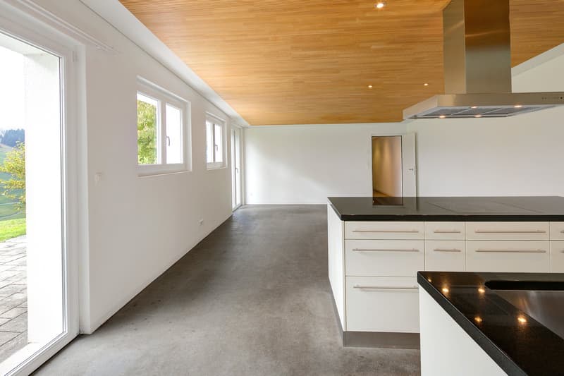 kitchen with large window, wooden ceiling, white walls, grey floor, black countertops, white cabinets, stainless steel kitchen hood, kitchen sink, under cabinet lighting