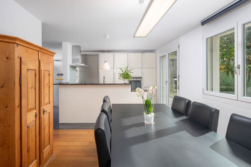 Dining area with a wooden floor, black dining table, white kitchen with appliances, sliding doors, wooden cabinet, flowers in a white vase