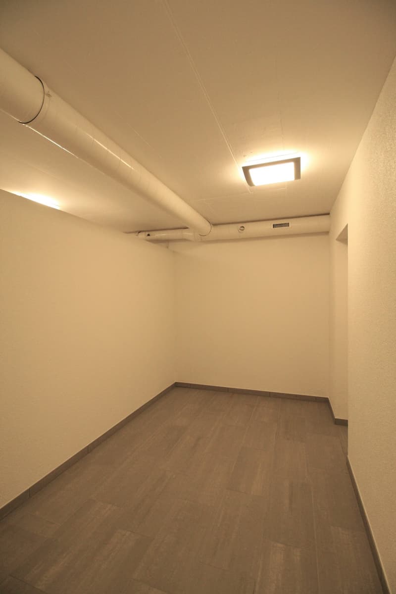 Empty room with wooden floors and white walls. There is a square ceiling light and pipework on the ceiling.