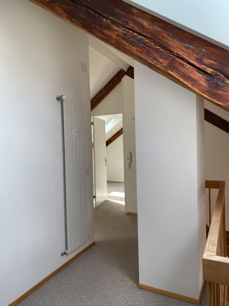 The image shows the interior of a room with exposed wooden beams on the ceiling, white walls, and a carpeted floor. There is a doorway leading to another room or area visible in the background.