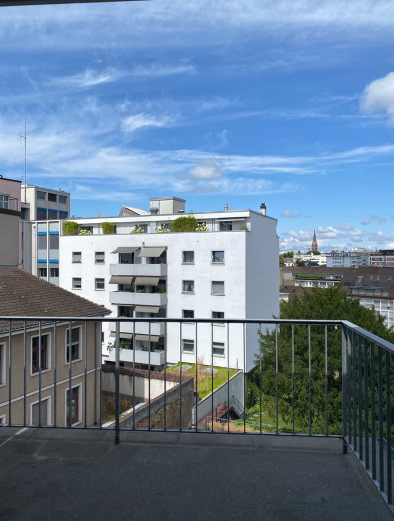 Multi-story apartment building with white exterior, balconies, and greenery on the rooftop. The building is surrounded by other urban structures and has a view of the city skyline.