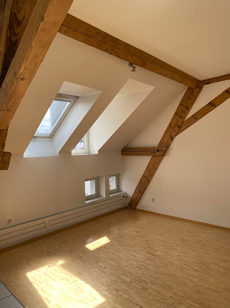 Attic bedroom with wooden beams, slanted ceilings, and skylights. The room has a hardwood floor and appears to be bright and airy.