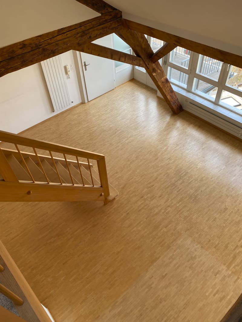 Spacious open-plan living area with wooden beams, large windows, and hardwood flooring. The room has a balcony or terrace visible.