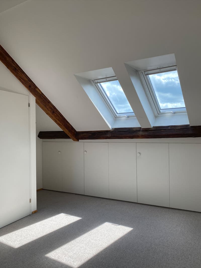 Spacious attic room with white walls, wooden beams, and two large skylights providing natural light. The room has built-in storage cabinets along one wall.