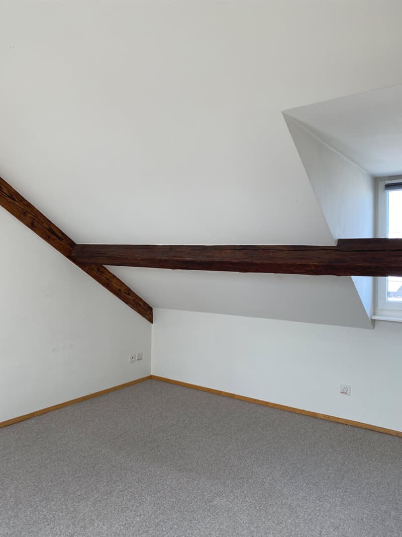 Attic or loft space with white walls, exposed wooden beams, and gray carpeted floor.