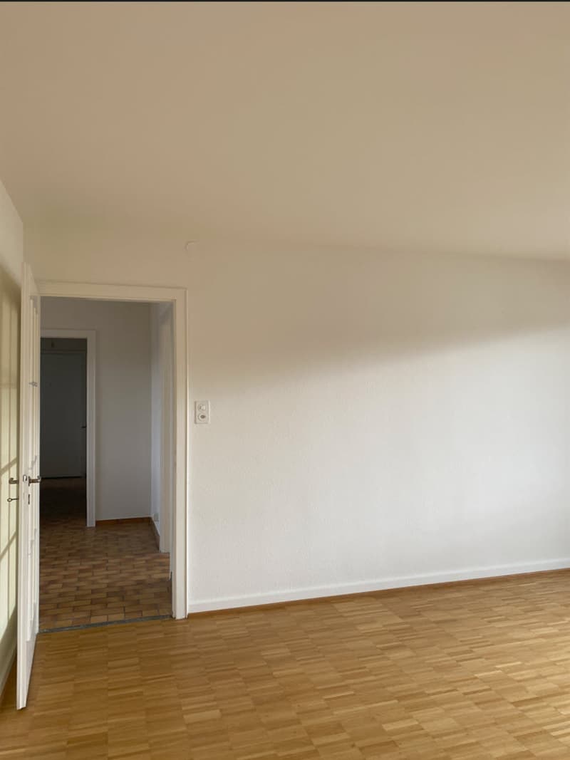 Empty room with wooden floor, white walls, open door, and another room visible in the background.