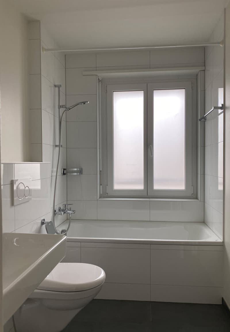 The image shows a modern and well-designed bathroom. The room features a large window that provides natural light, a bathtub, and a toilet. The walls are tiled in a neutral color scheme, creating a clean and minimalist aesthetic. The bathroom appears to b