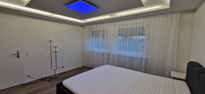 Spacious bedroom with a large window, white curtains, and a blue-lit ceiling. The room has a king-size bed, bedside tables, and a floor lamp.