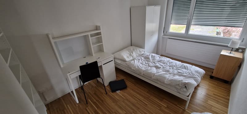 Spacious bedroom with hardwood flooring, white walls, a bed with white bedding, a wardrobe, and a desk with a chair. The room has a large window providing natural light.