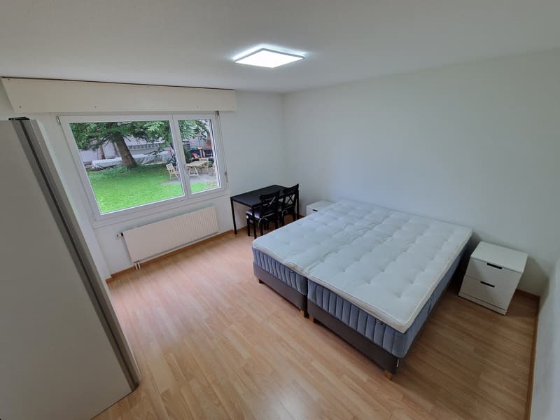 Room with wooden floor, white walls, large window with outdoor view, double bed, white nightstands, desk with chairs, and ceiling light.