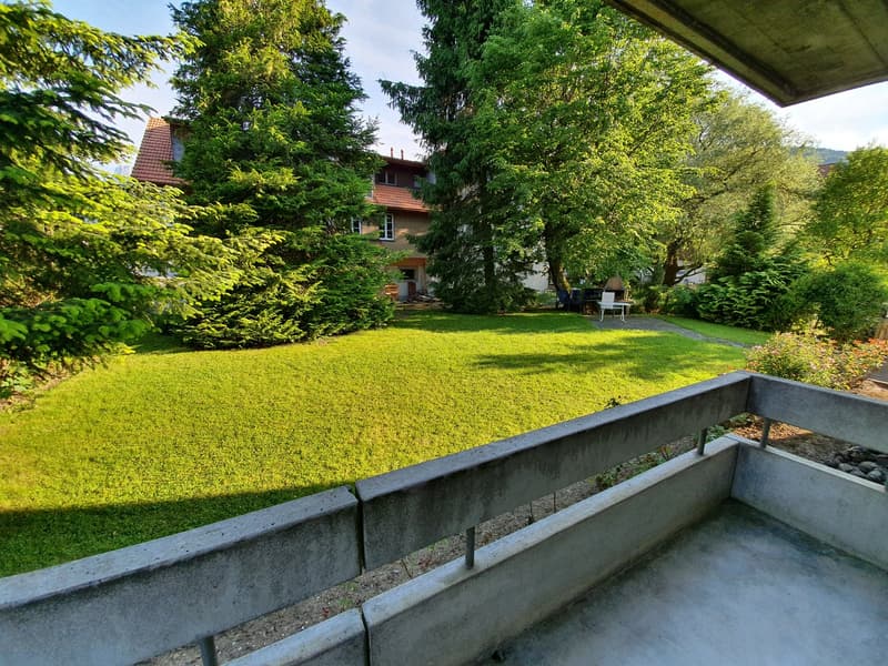 Balcony overlooking a well-maintained garden with a lush lawn and various trees and plants.
