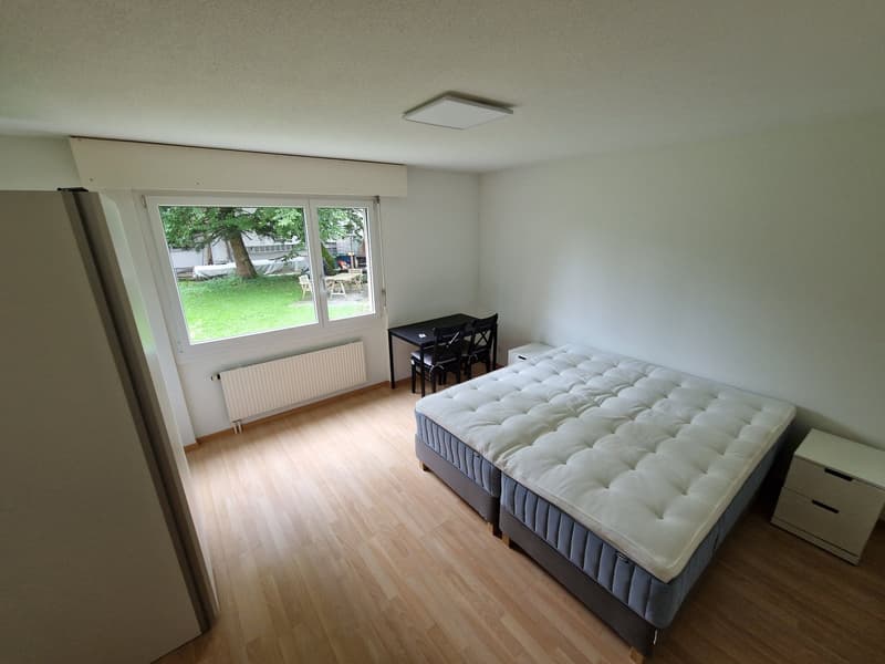 a room with a double bed, black desk and chairs, white wardrobe, wooden floor, large window, white walls, heating panel